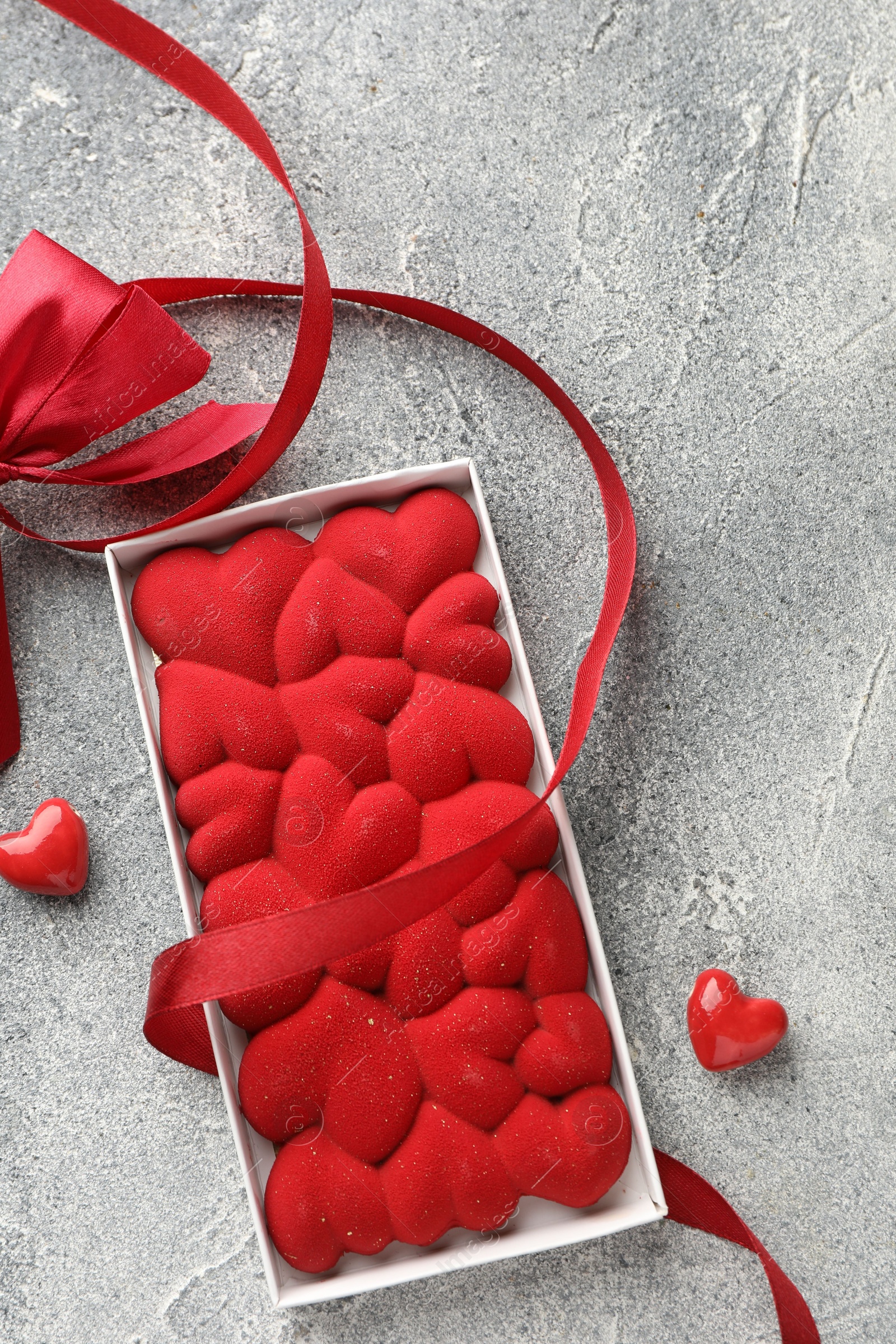 Photo of Chocolate bar decorated with hearts, candies and ribbon on grey textured background, top view