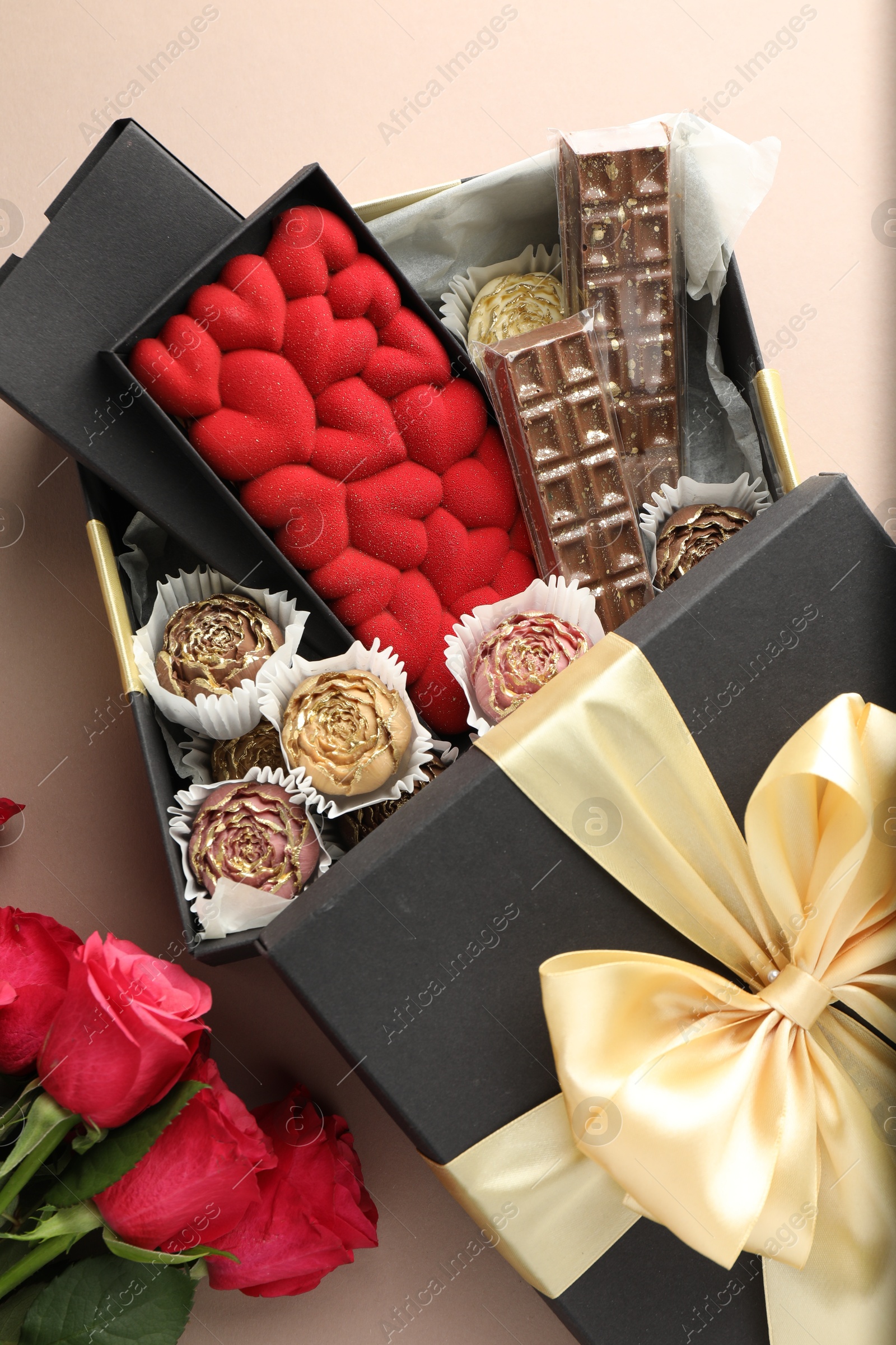 Photo of Different delicious chocolate sweets in box and roses on beige background, top view