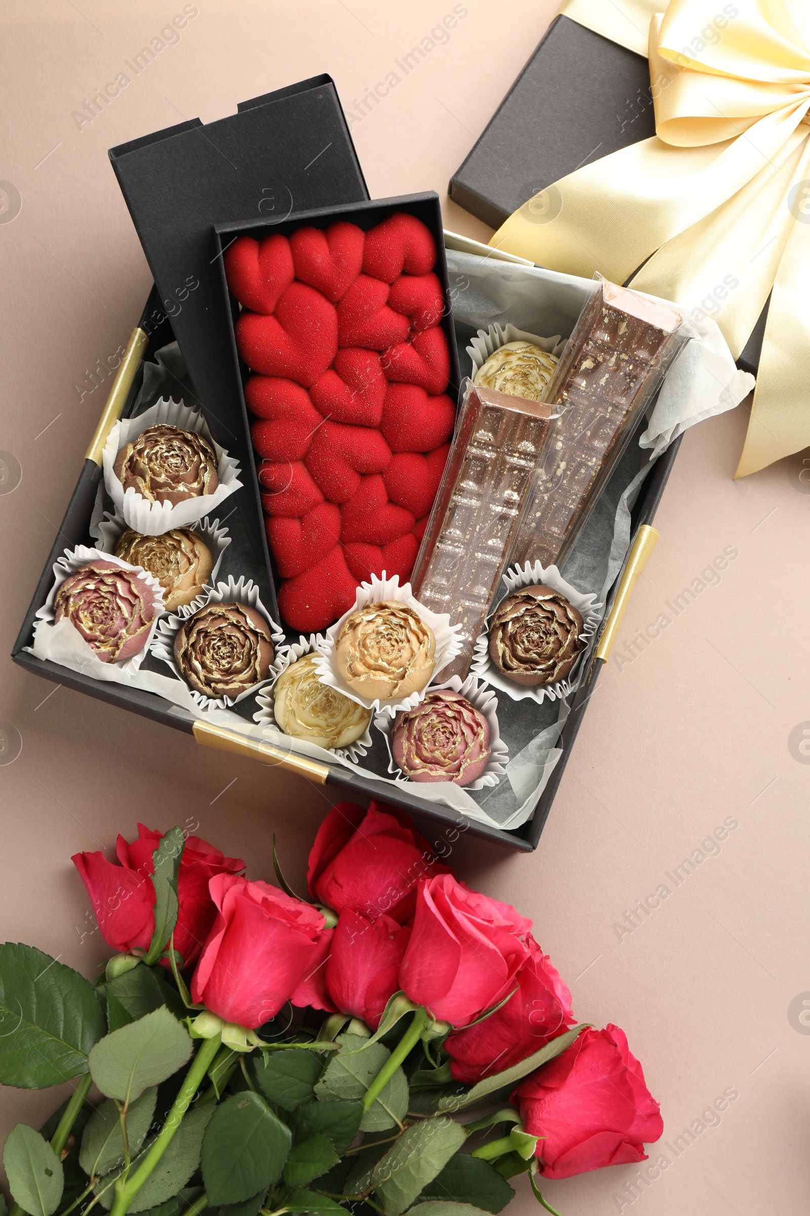 Photo of Different delicious chocolate sweets in box and roses on beige background, top view