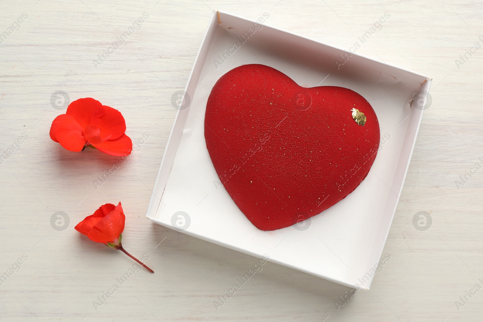 Photo of Heart shaped chocolate in box and flowers on light wooden background, top view