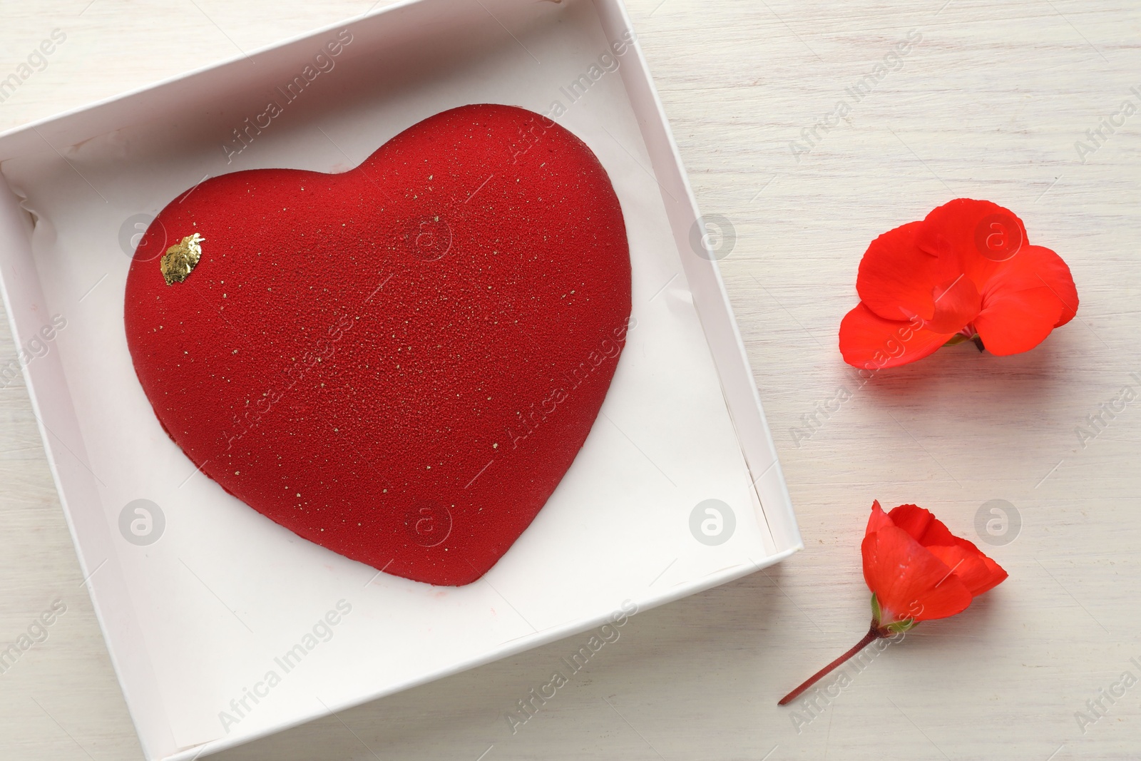 Photo of Heart shaped chocolate in box and flowers on light wooden background, top view