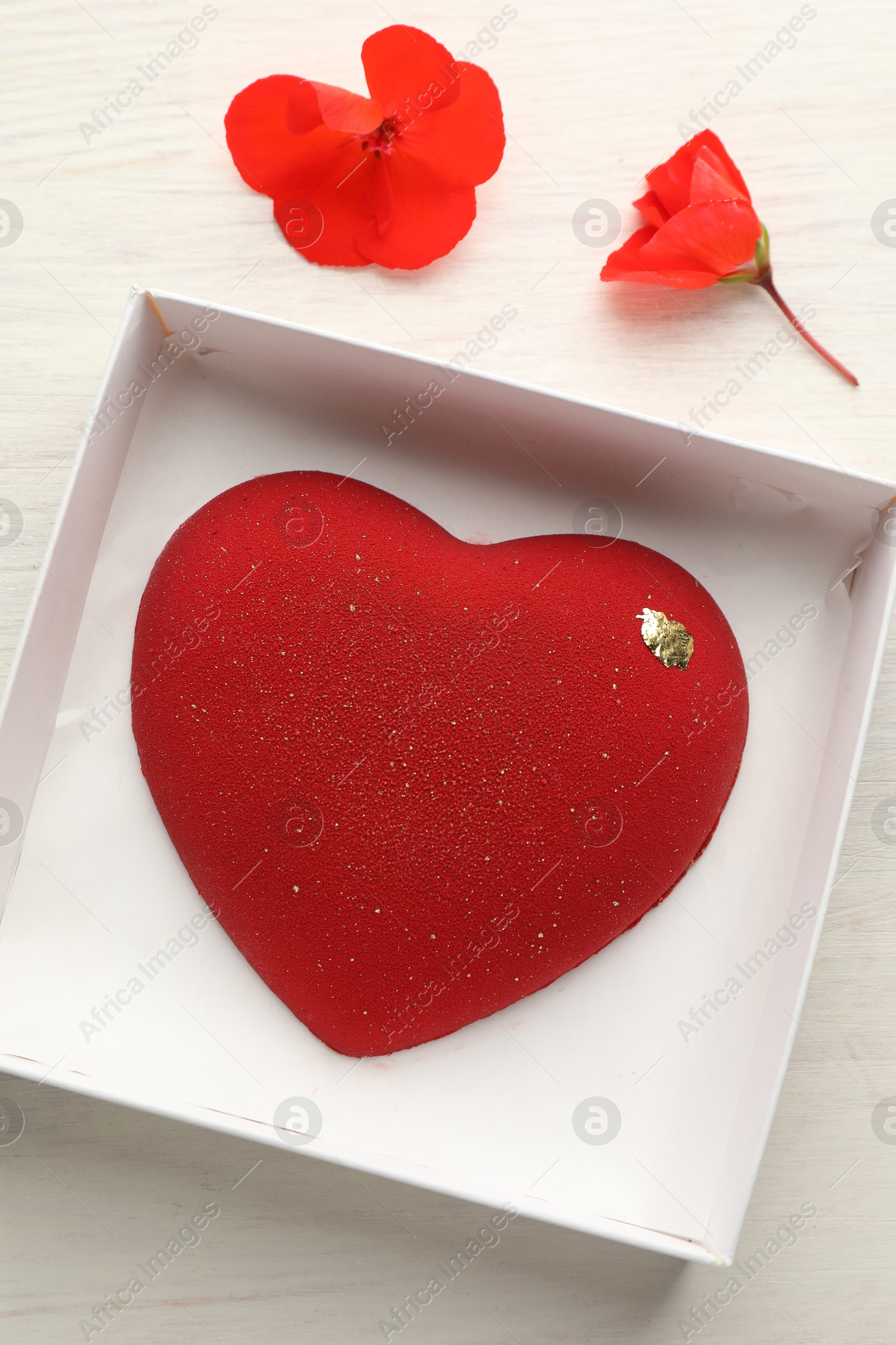 Photo of Heart shaped chocolate in box and flowers on light wooden background, top view