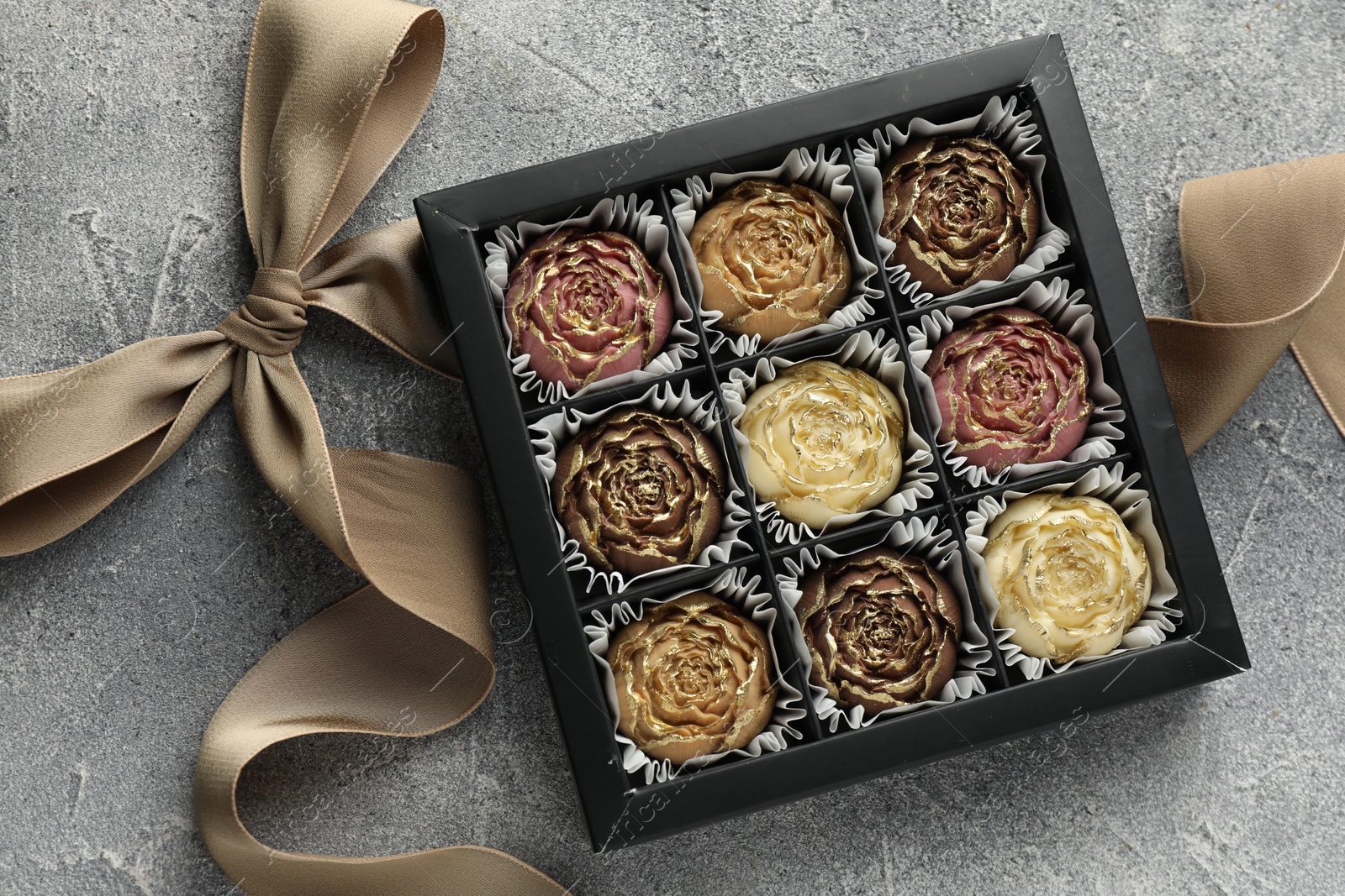 Photo of Delicious flower shaped chocolate bonbons in box and bow on grey textured background, top view