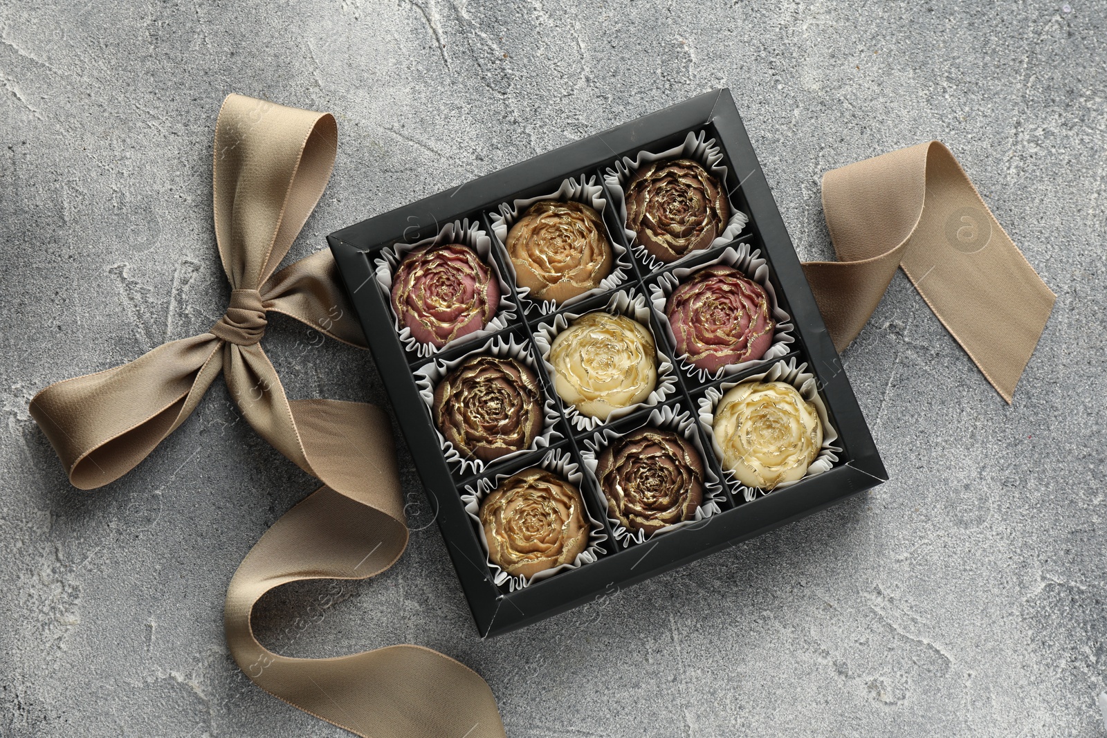 Photo of Delicious flower shaped chocolate bonbons in box and bow on grey textured background, top view