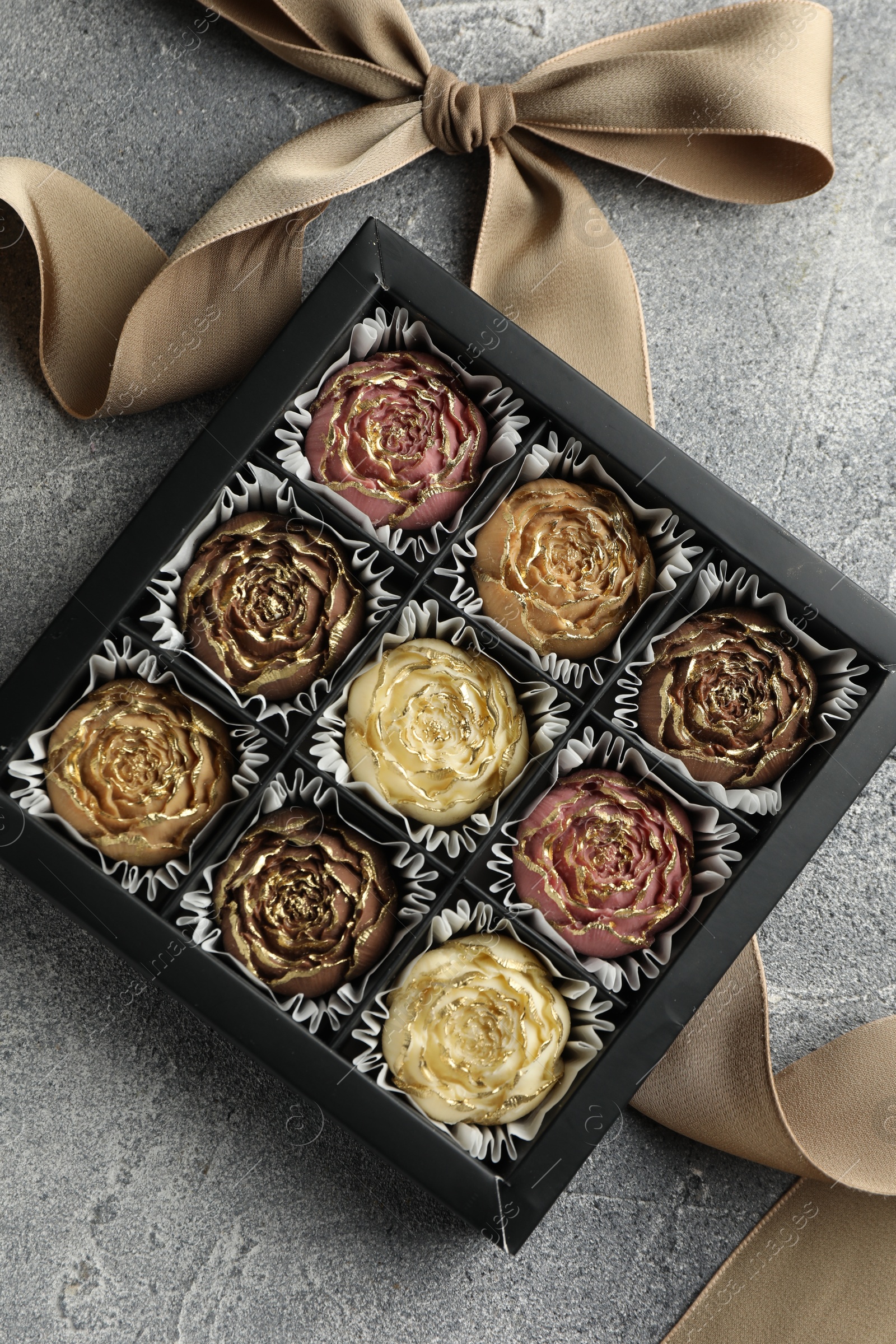 Photo of Delicious flower shaped chocolate bonbons in box and bow on grey textured background, top view