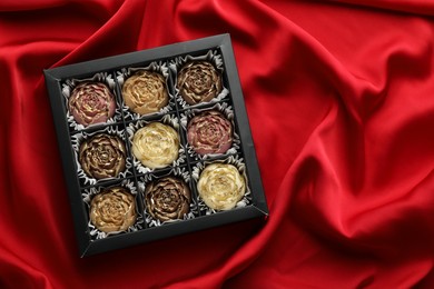 Photo of Delicious flower shaped chocolate bonbons in box on red fabric background, top view. Space for text