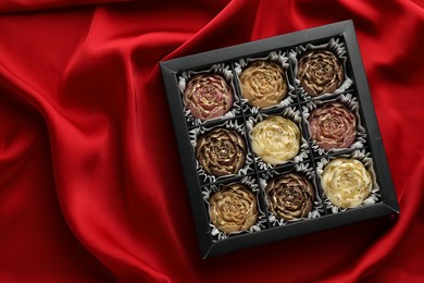 Photo of Delicious flower shaped chocolate bonbons in box on red fabric background, top view. Space for text