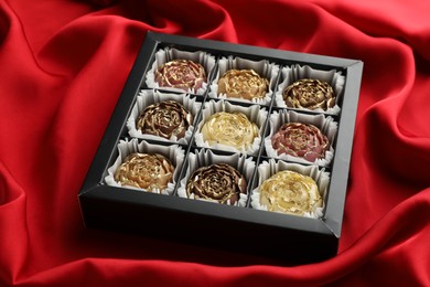 Photo of Delicious flower shaped chocolate bonbons in box on red fabric background, closeup