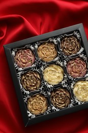 Photo of Delicious flower shaped chocolate bonbons in box on red fabric background, top view