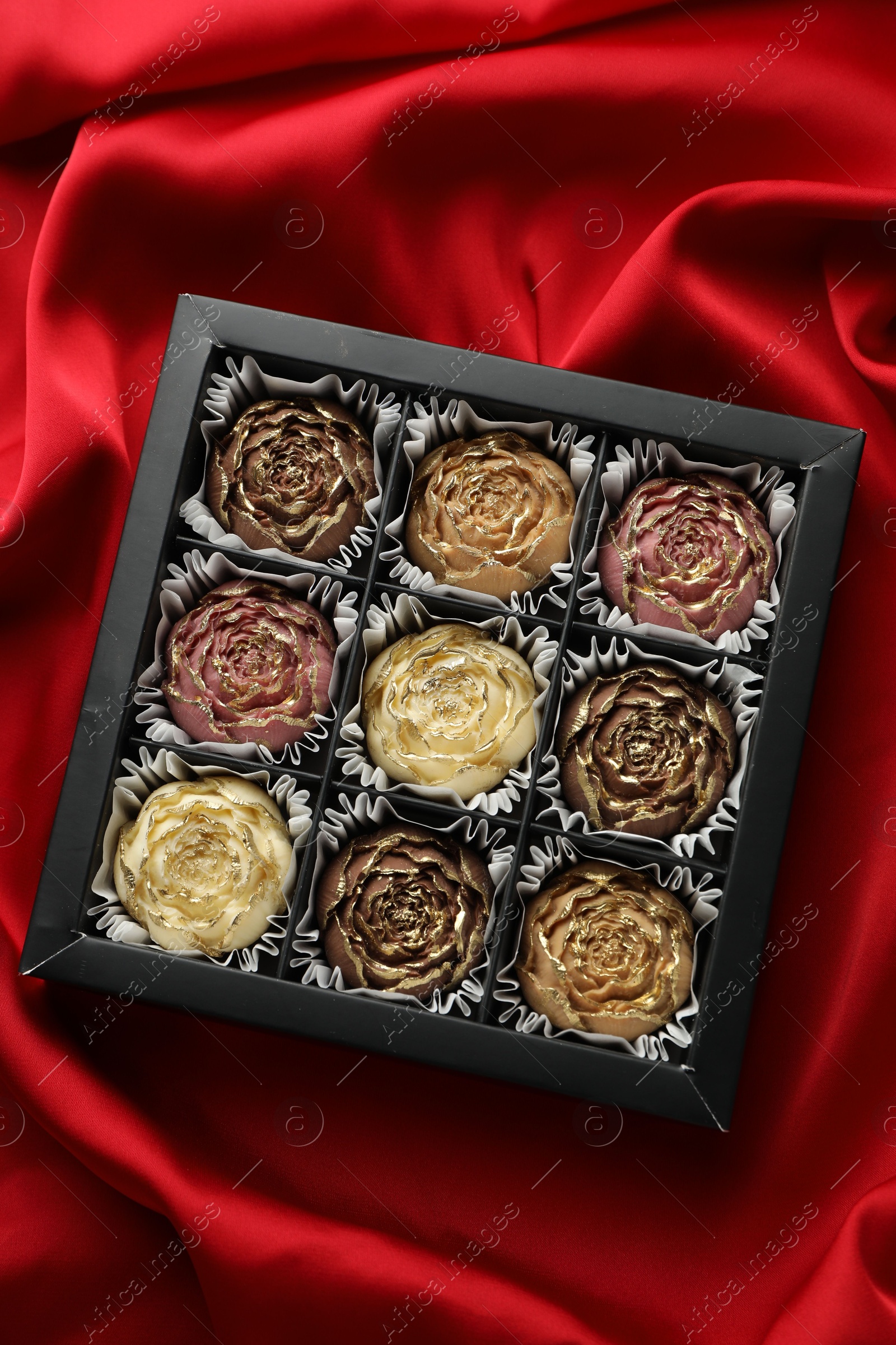 Photo of Delicious flower shaped chocolate bonbons in box on red fabric background, top view