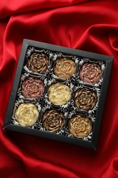 Photo of Delicious flower shaped chocolate bonbons in box on red fabric background, top view