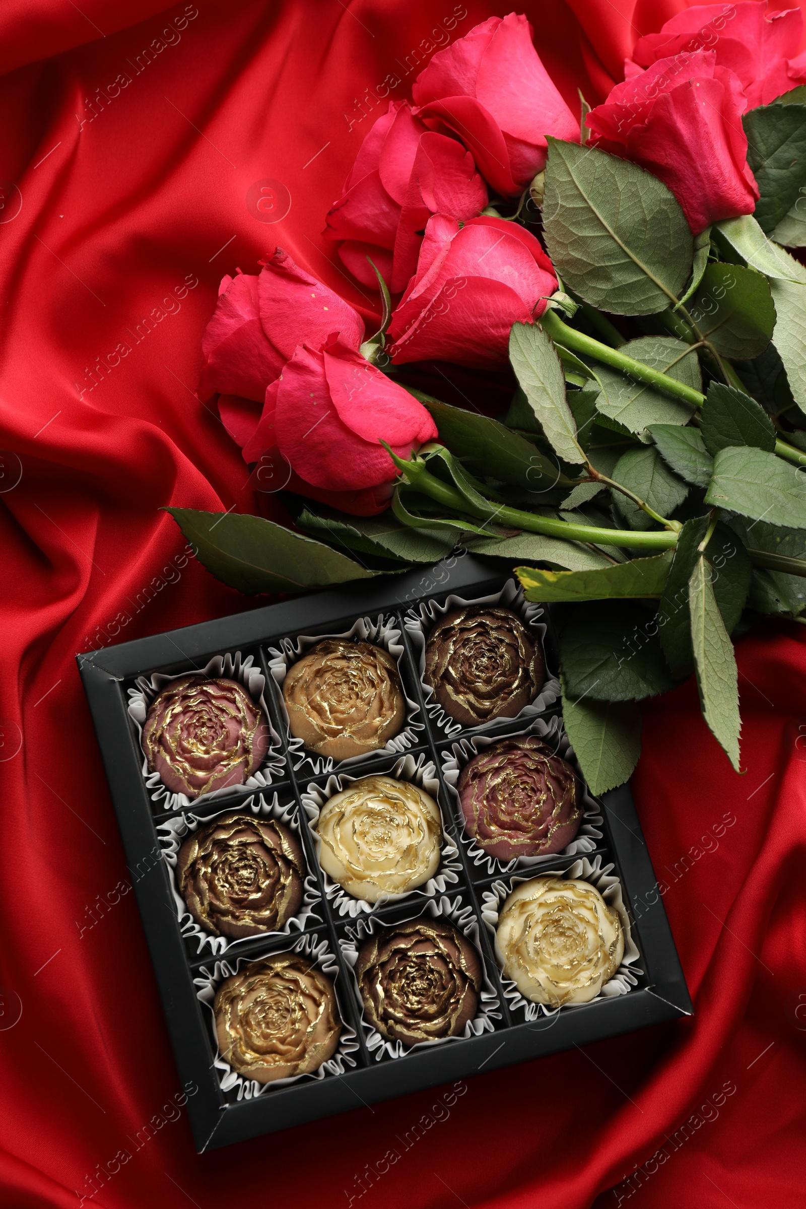 Photo of Delicious flower shaped chocolate bonbons in box and roses on red fabric background, top view