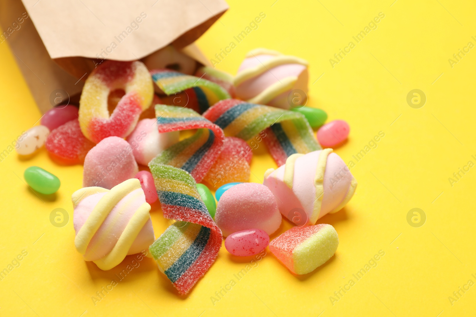 Photo of Paper bag with many different tasty candies and marshmallows on yellow background, closeup