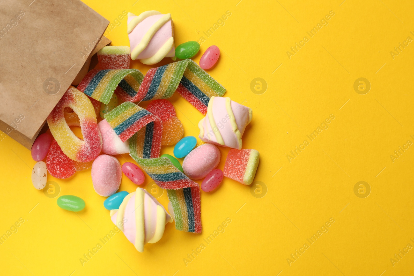 Photo of Paper bag with many different tasty candies and marshmallows on yellow background, flat lay. Space for text