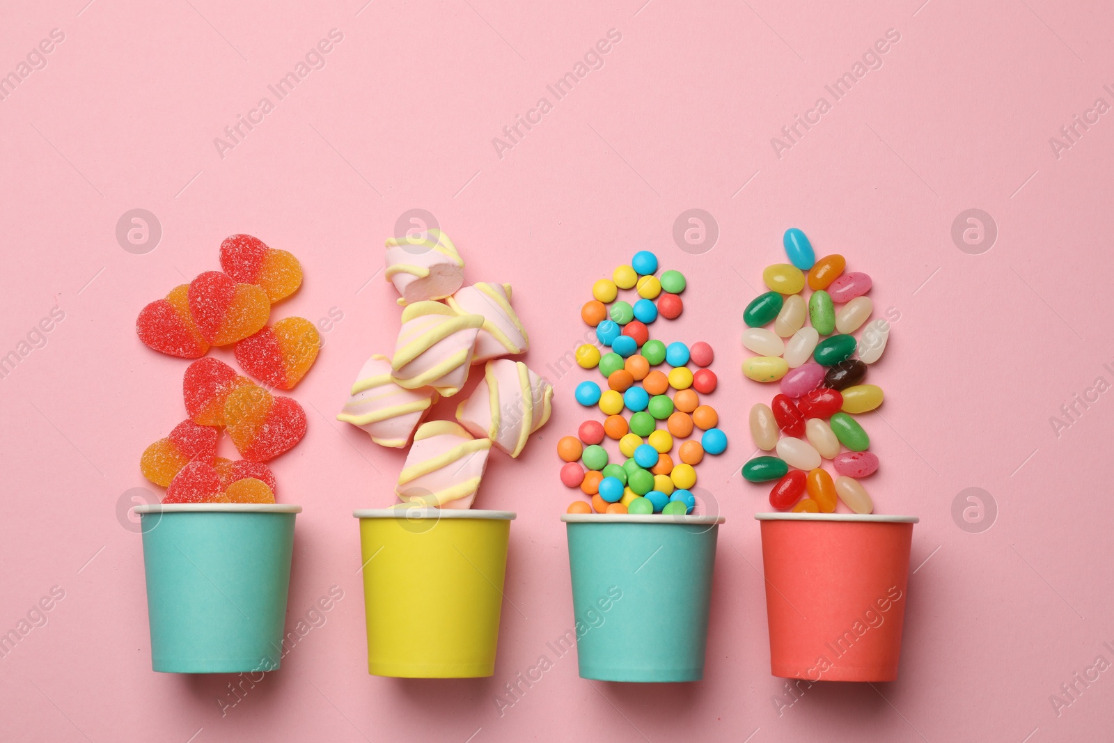 Photo of Tasty candies, marshmallows and paper cups on pink background, flat lay