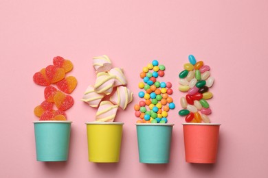 Photo of Tasty candies, marshmallows and paper cups on pink background, flat lay