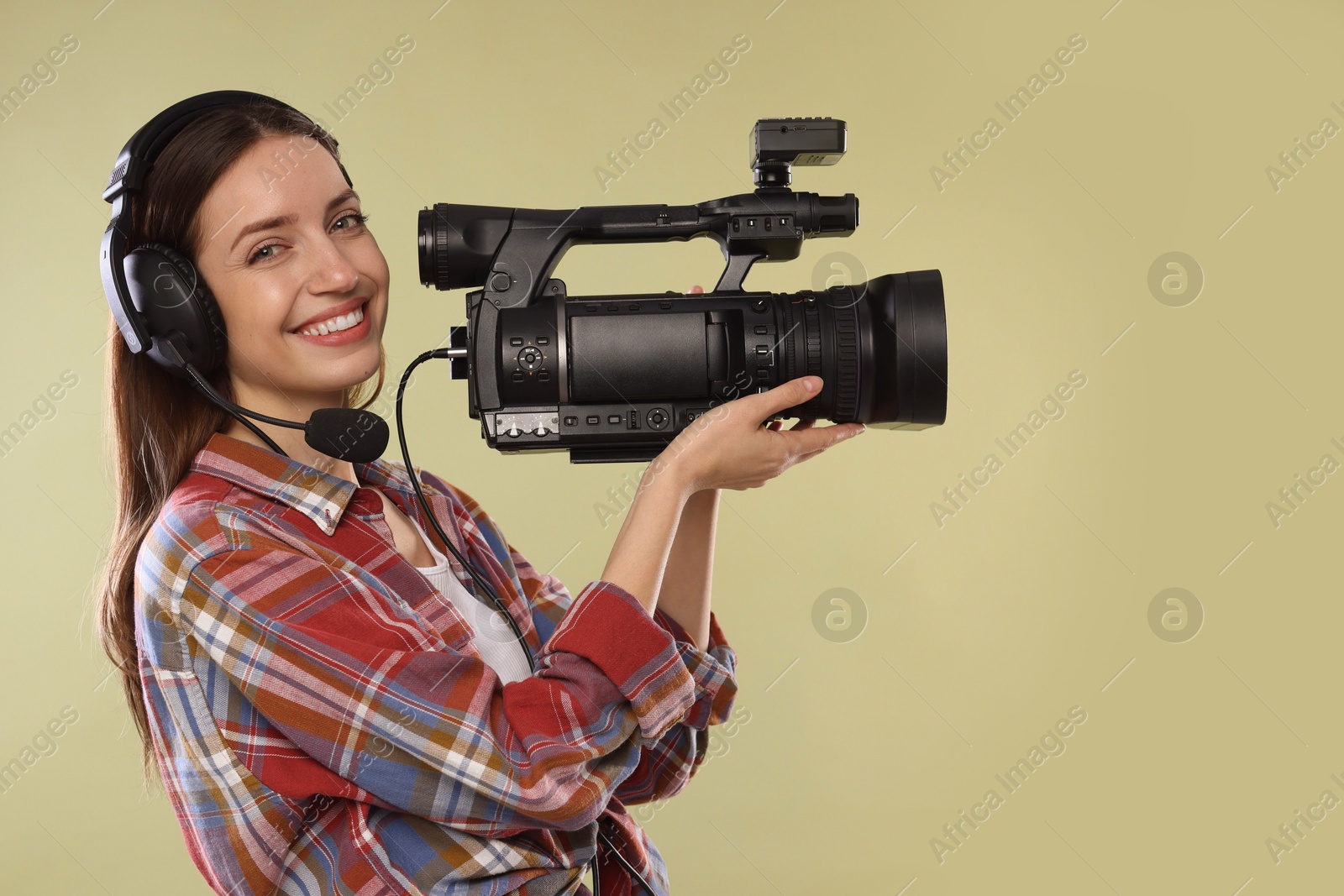 Photo of Happy woman with professional video camera and headset on pale olive background, space for text