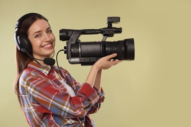 Happy woman with professional video camera and headset on pale olive background, space for text