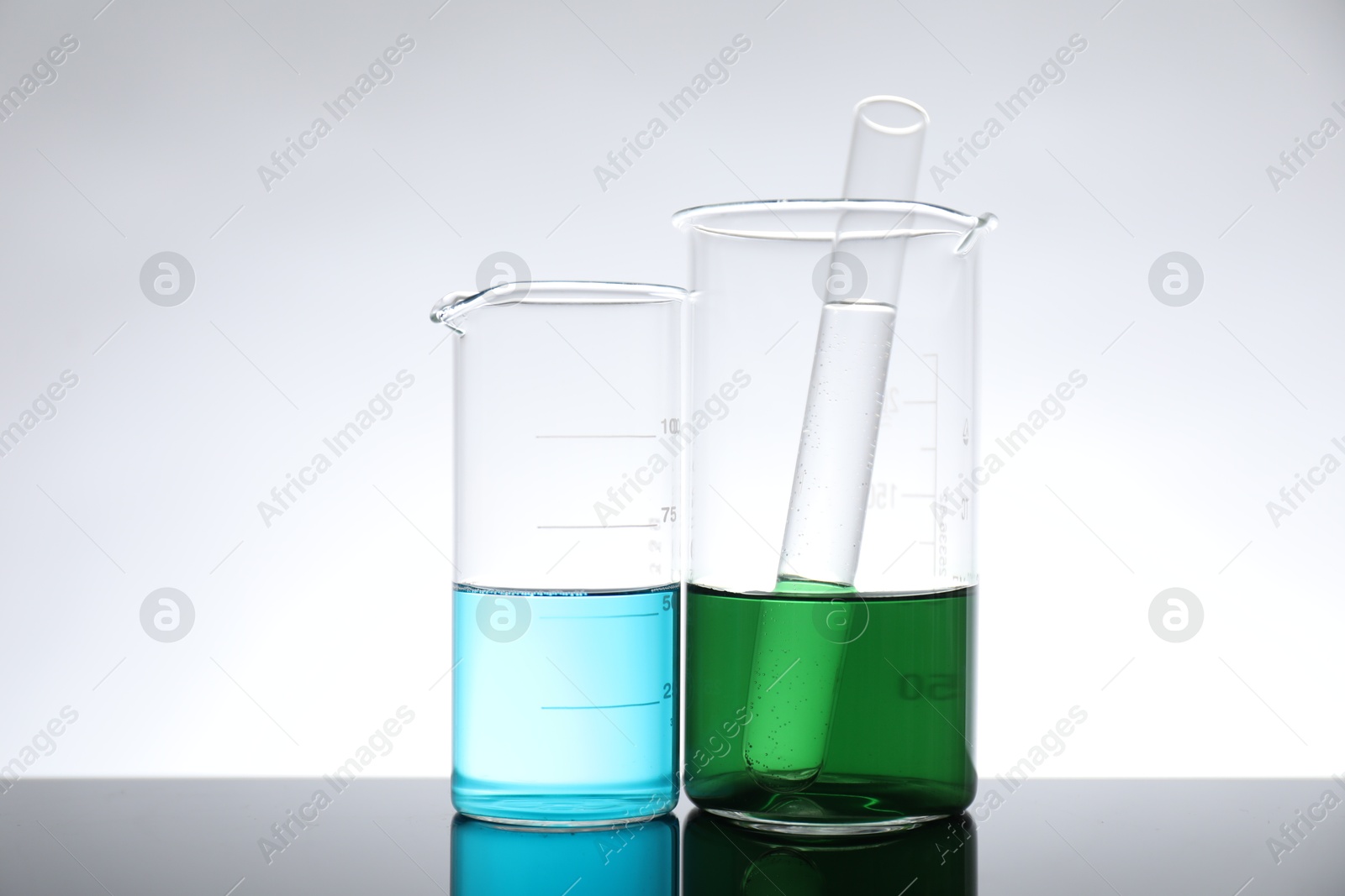 Photo of Beakers and test tube with liquid on mirror surface against light background. Laboratory glassware