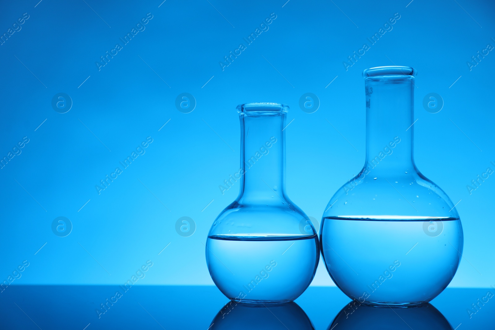 Photo of Flasks with liquid on mirror surface against light blue background, space for text. Laboratory equipment