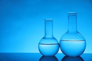 Photo of Flasks with liquid on mirror surface against light blue background, space for text. Laboratory equipment