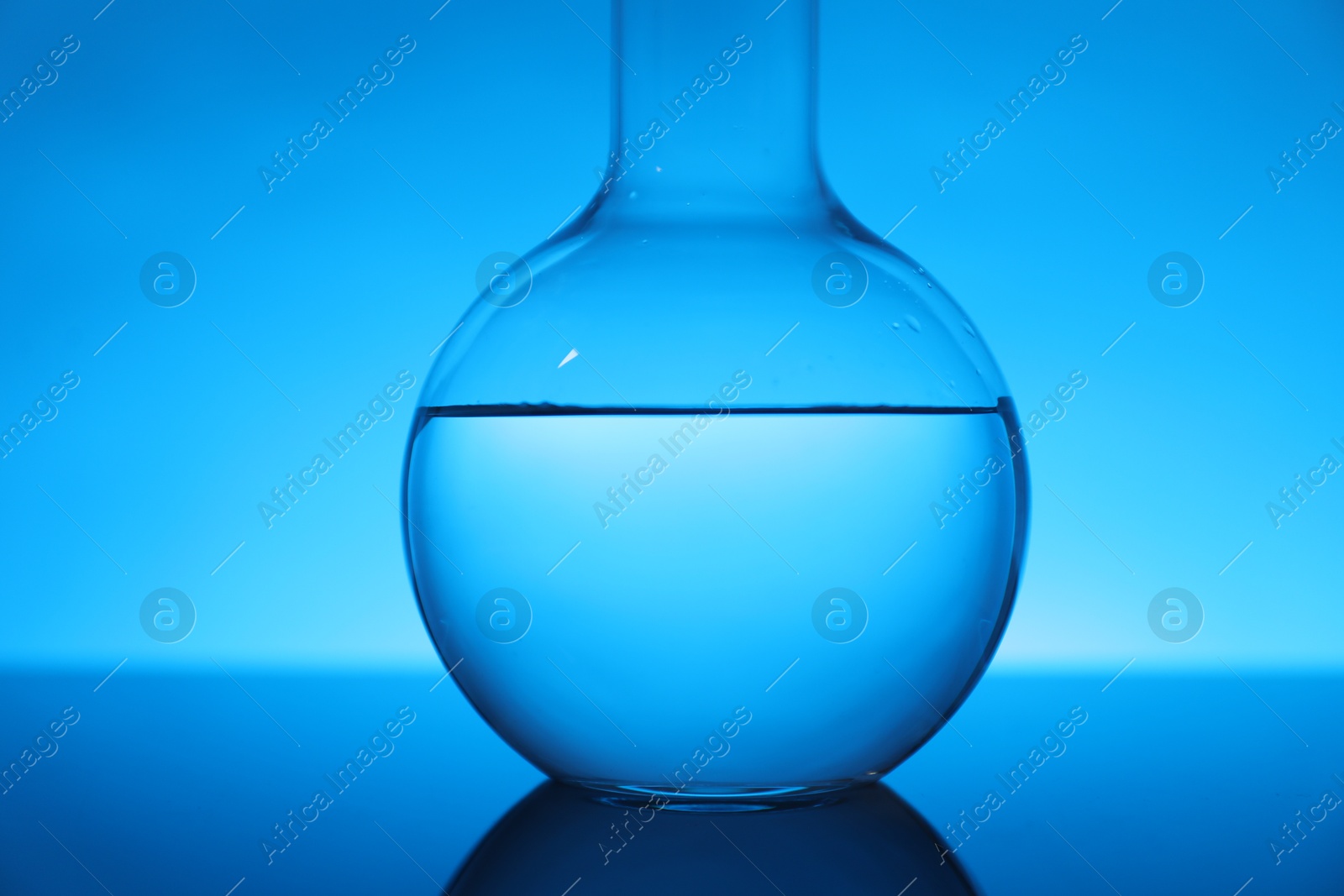 Photo of Flask with liquid on mirror surface against light blue background, closeup. Laboratory equipment