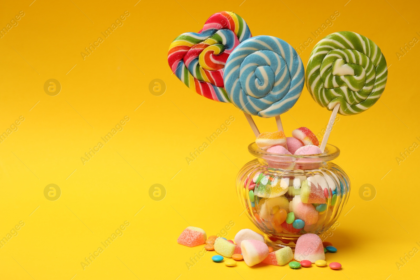 Photo of Different tasty candies in glass jar on yellow background. Space for text