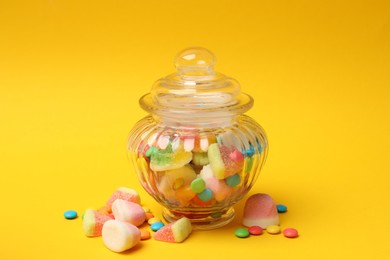 Photo of Different tasty candies in glass jar on yellow background, closeup