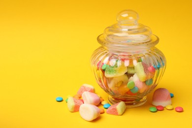 Photo of Different tasty candies in glass jar on yellow background, closeup. Space for text