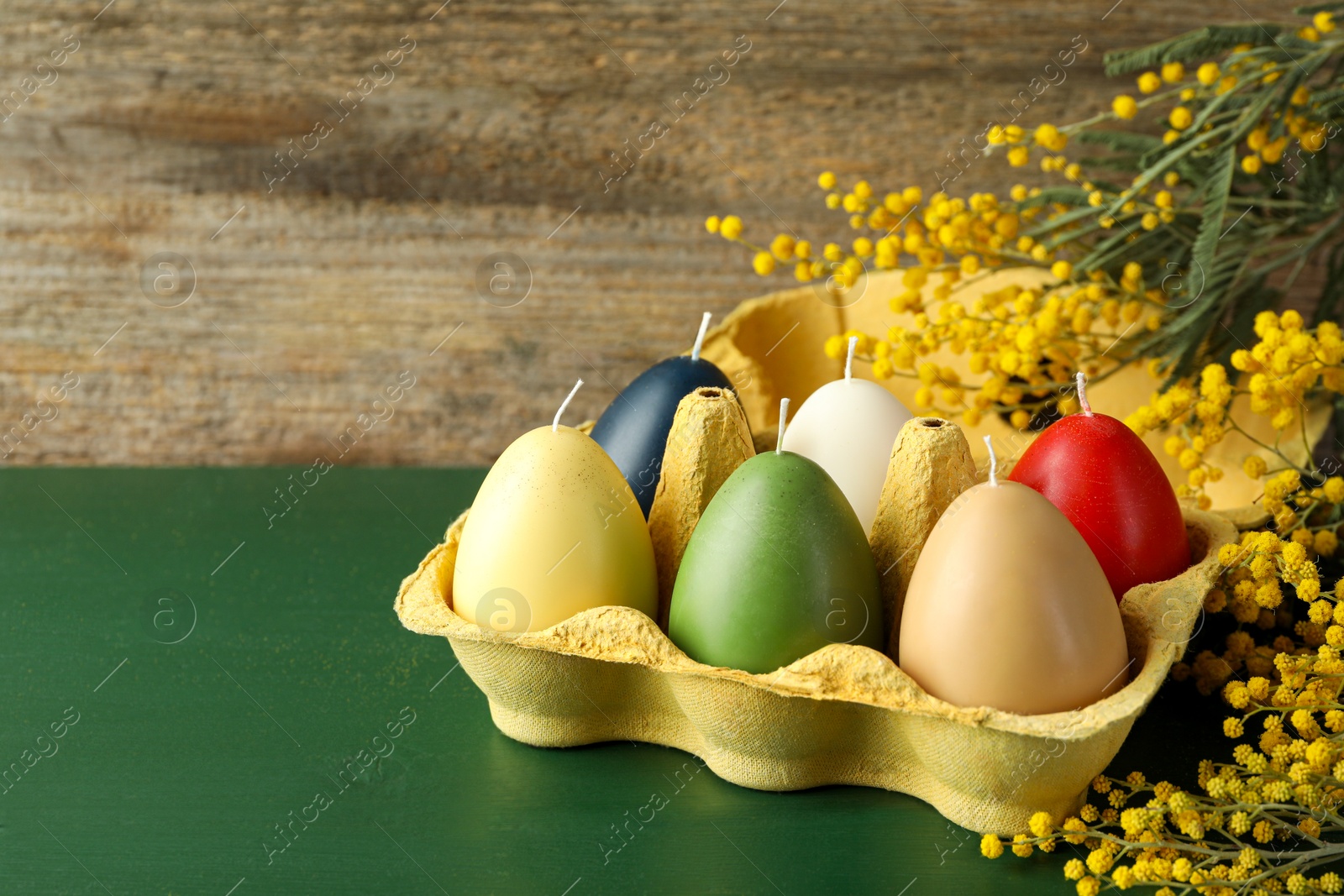 Photo of Colorful egg-shaped candles in carton and mimosa flowers on green wooden table, closeup with space for text. Easter decor