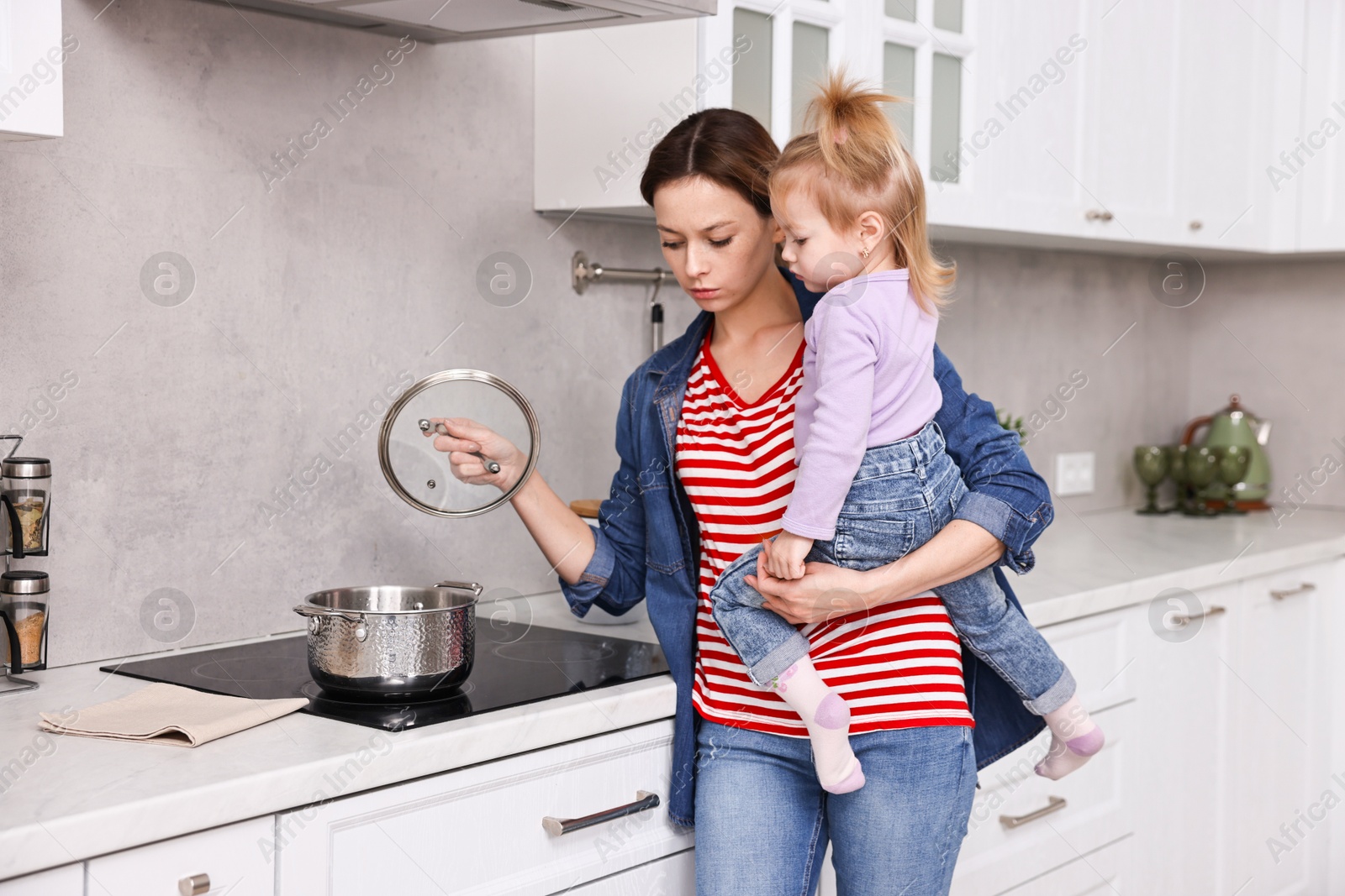 Photo of Busy housewife cooking and holding her little daughter in kitchen