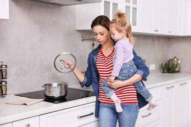 Busy housewife cooking and holding her little daughter in kitchen