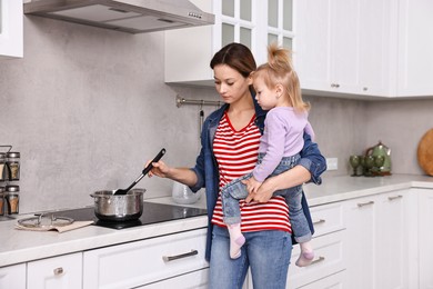 Busy housewife cooking and holding her little daughter in kitchen