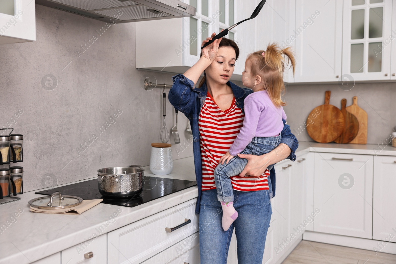 Photo of Tired housewife cooking and holding her little daughter in kitchen