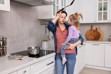 Tired housewife cooking and holding her little daughter in kitchen