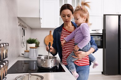 Photo of Busy housewife cooking and holding her little daughter in kitchen