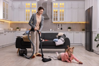 Photo of Smiling housewife cleaning floor with vacuum cleaner while her little daughter playing at home