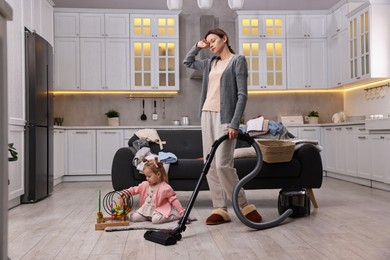 Tired housewife cleaning floor with vacuum cleaner while her little daughter playing at home