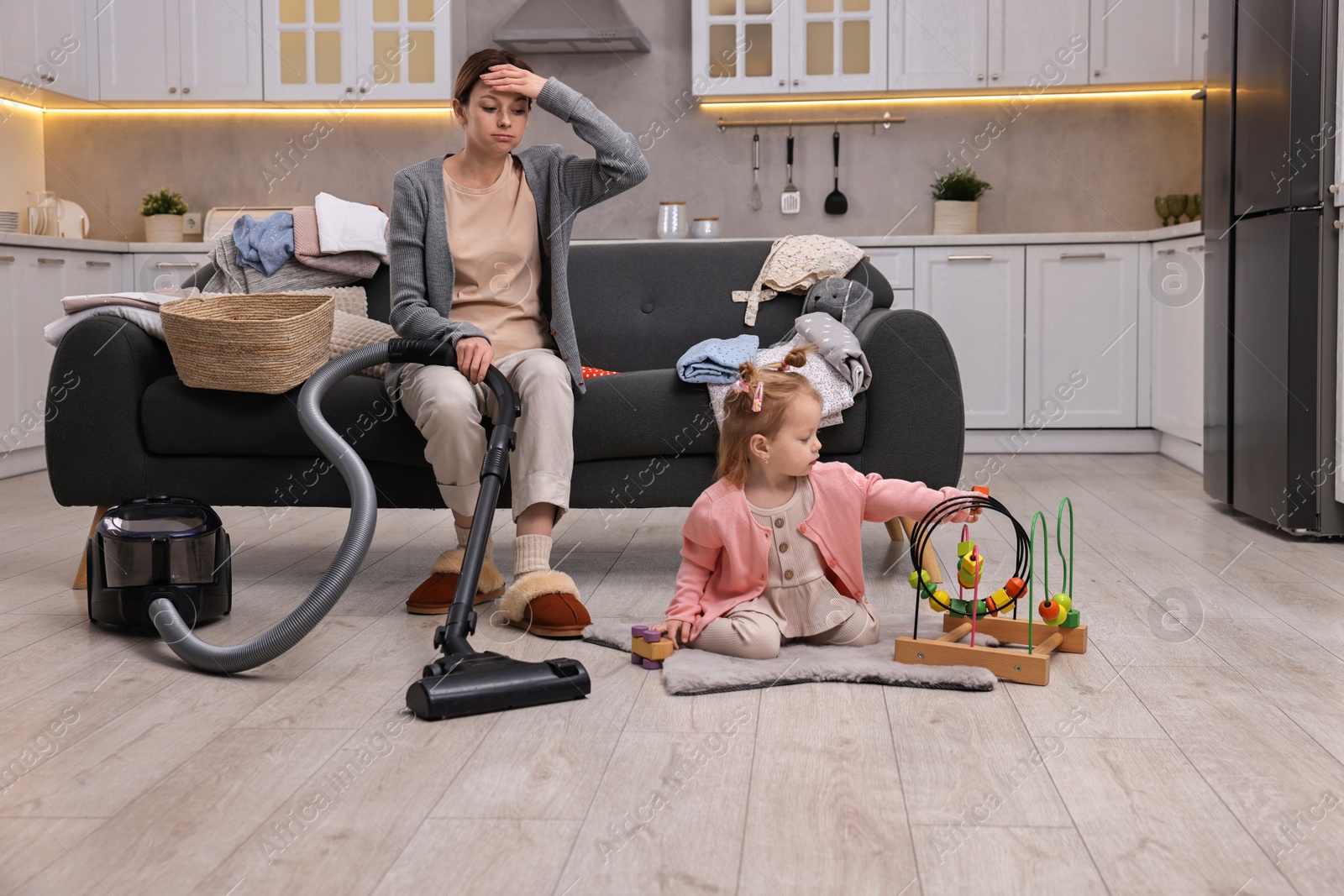 Photo of Tired housewife with vacuum cleaner sitting among messy laundry on sofa while her little daughter playing at home