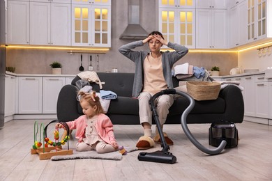 Photo of Tired housewife with vacuum cleaner sitting among messy laundry on sofa while her little daughter playing at home