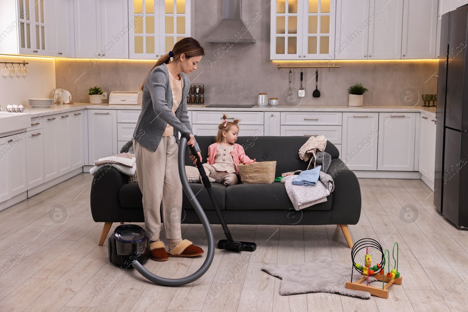 Photo of Housewife cleaning floor with vacuum cleaner near sofa and her little daughter at home