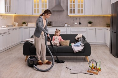 Photo of Housewife cleaning floor with vacuum cleaner near sofa and her little daughter at home