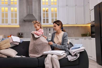 Housewife with her little daughter folding laundry on sofa at home