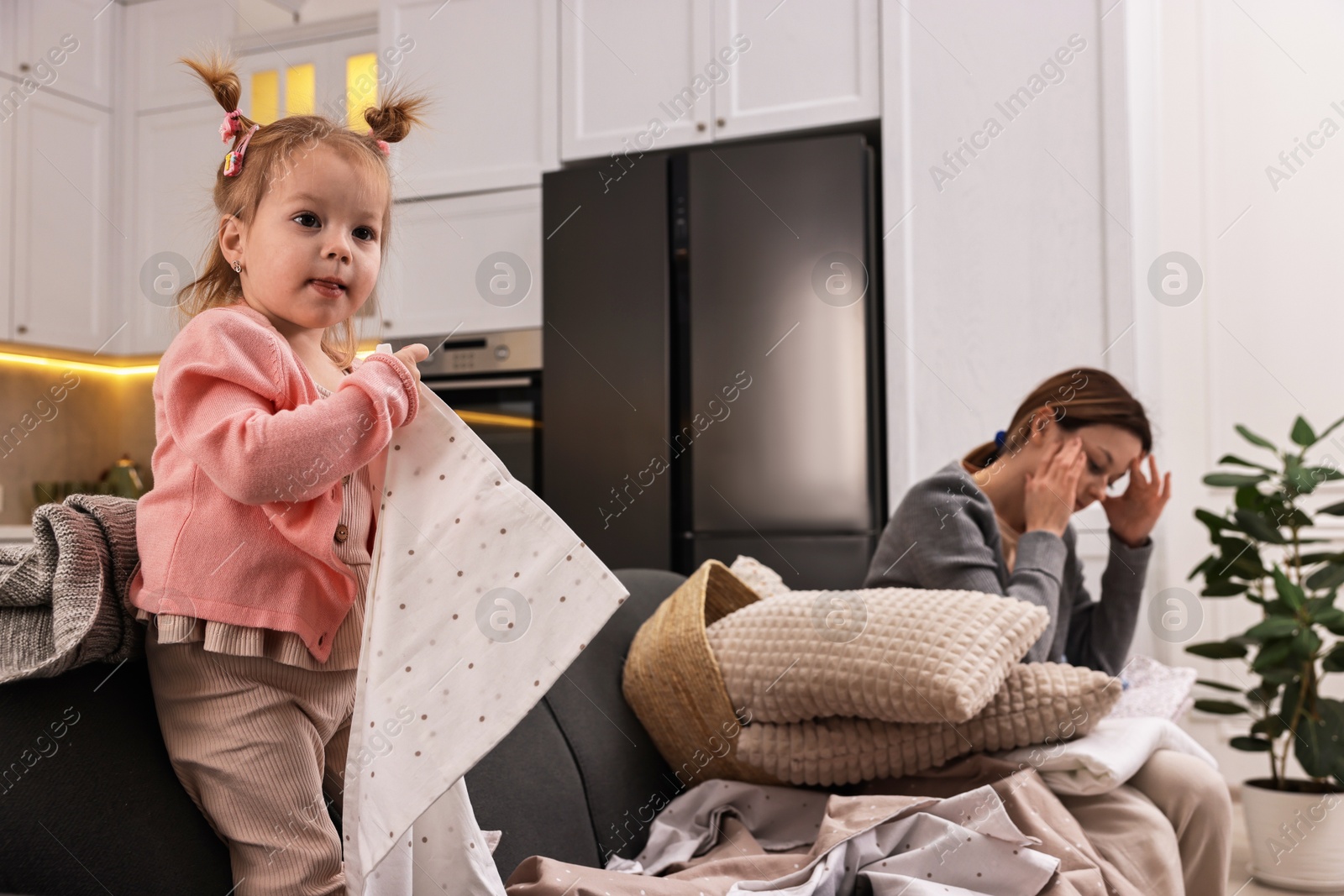 Photo of Tired housewife with her little daughter among messy laundry on sofa at home