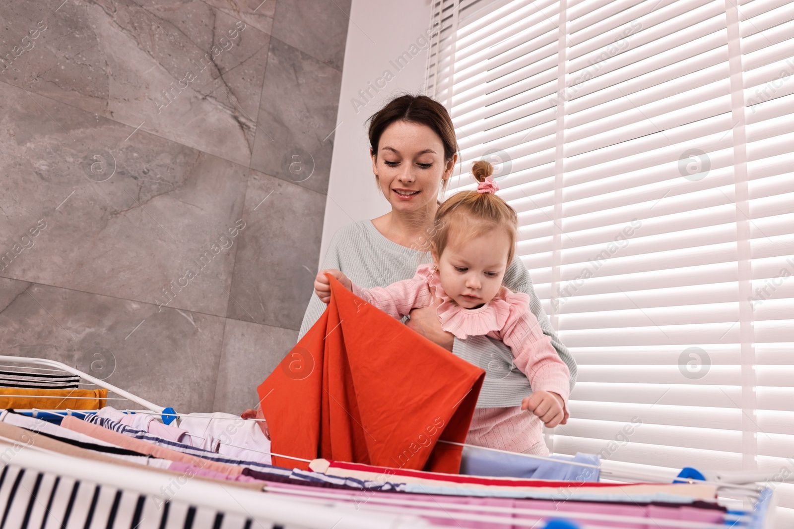 Photo of Smiling housewife with her daughter hanging clothes on drying rack at home, low angle view