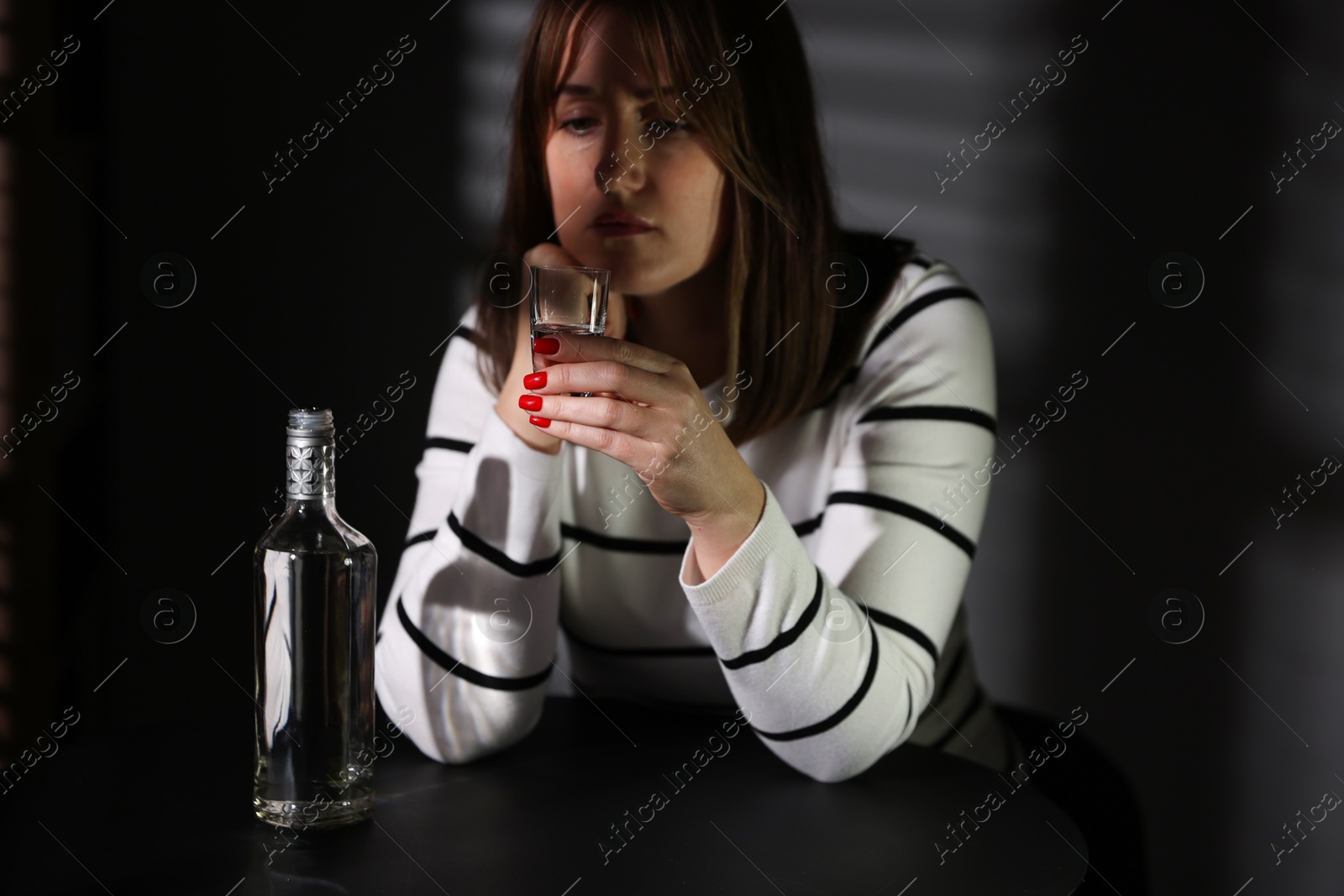 Photo of Alcohol addiction. Woman with shot glass of vodka and bottle at table indoors