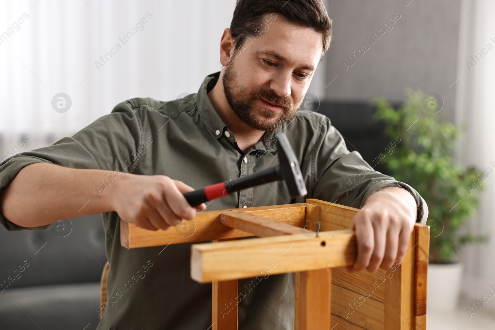 Photo of Man repairing wooden stool with nail and hammer at home