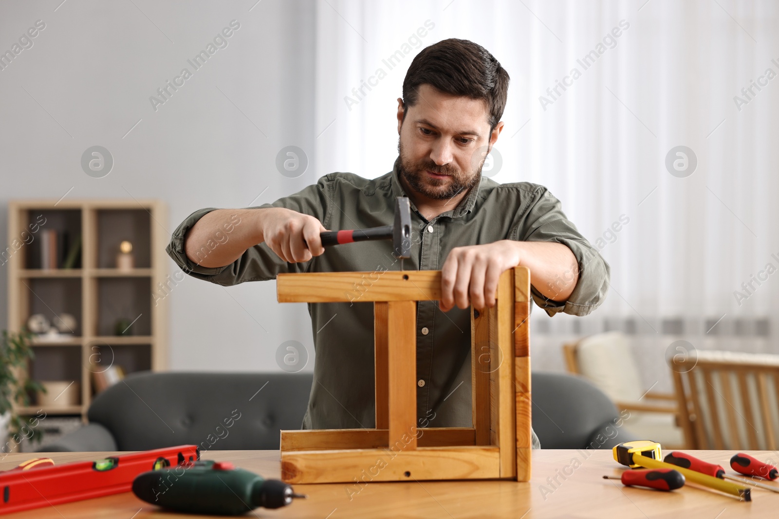 Photo of Man repairing wooden stool with nail and hammer at home