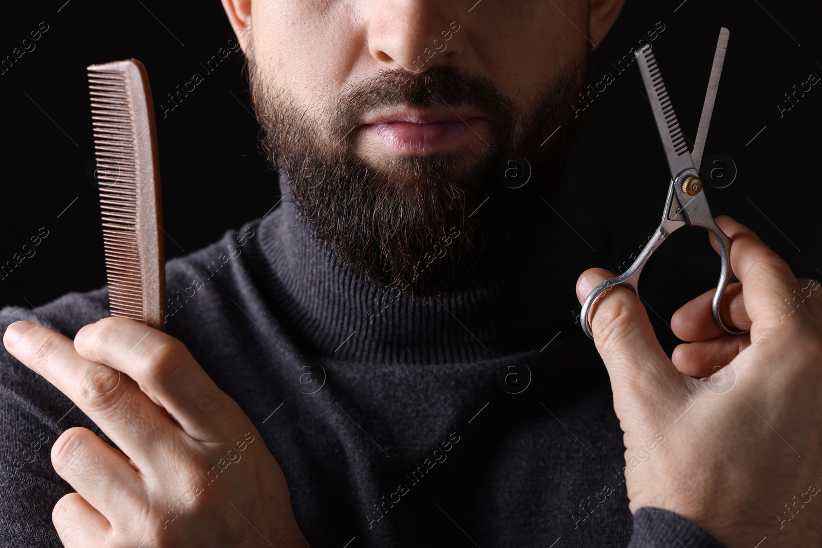 Photo of Bearded man holding comb and scissors on black background, closeup