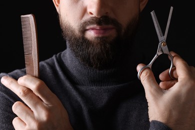 Photo of Bearded man holding comb and scissors on black background, closeup